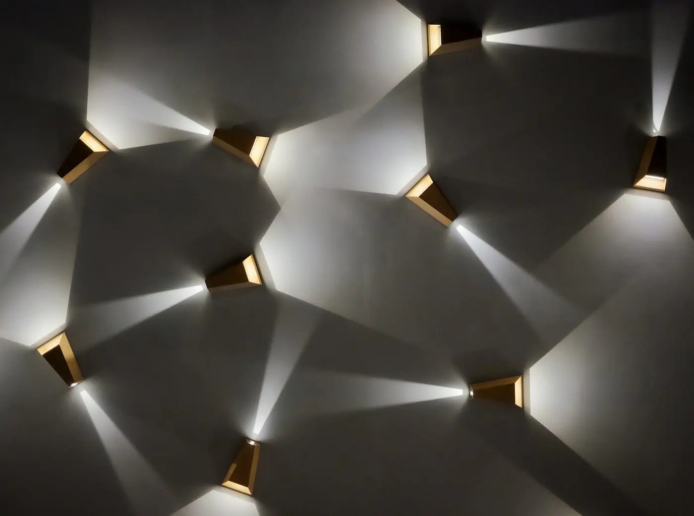 Angular gold-toned wall sconces casting beams of light and geometric shadows across a white wall